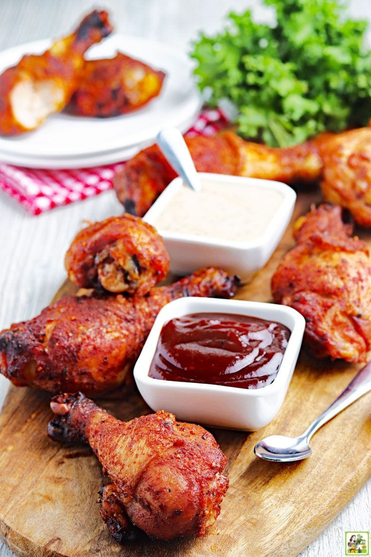 A board of BBQ chicken drumsticks with a plate in the background of chicken legs with bowls of BBQ sauce and Alabama BBQ sauce.