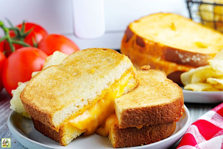Ψητά σάντουιτς τυριών φριτέζας αέρα σε ένα άσπρο πιάτο με μια κόκκινη πετσέτα και ντομάτες μπροστά από μια φριτέζα αέρα.