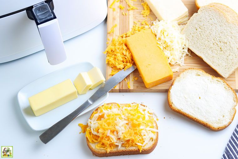 Εξοπλισμός και συστατικά που απαιτούνται για την παρασκευή ενός σάντουιτς ψητού τυριού με φριτέζα αέρα, όπως βούτυρο, ψωμί, τριμμένο τυρί, μαχαίρι και φριτέζα αέρα.