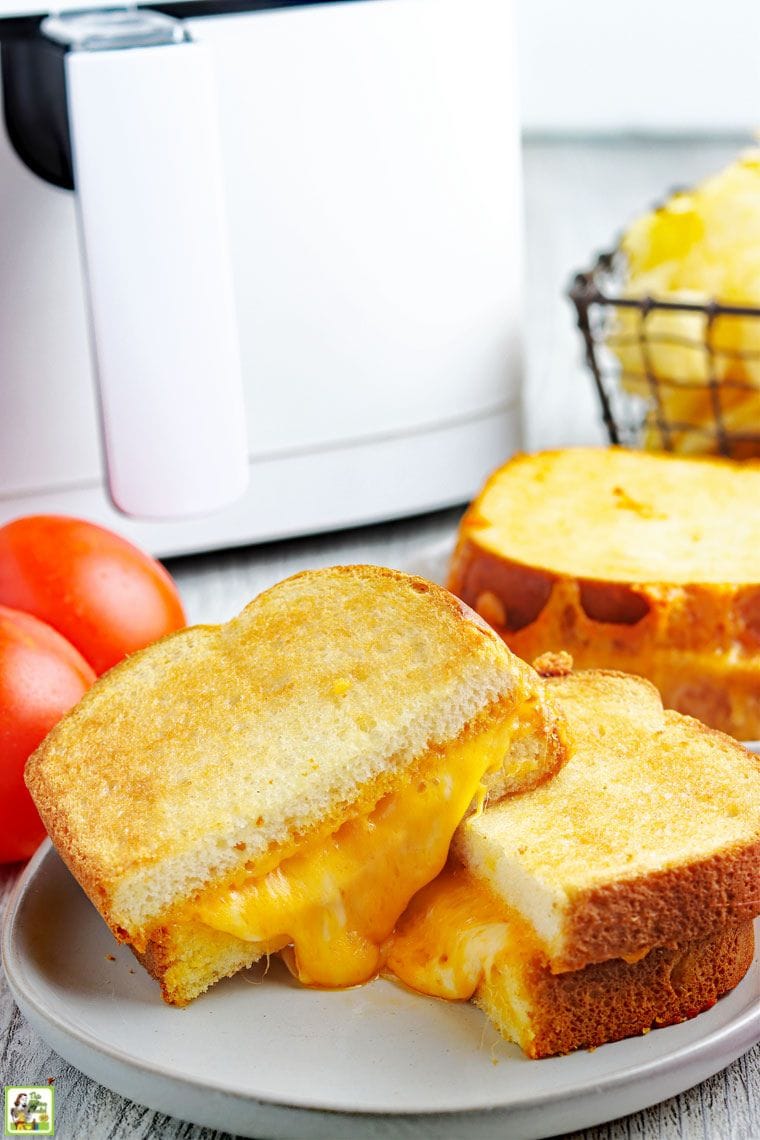 Sliced melted grill cheese sandwich on a plate in front of an air fryer with tomatoes and another grill cheese sandwich.