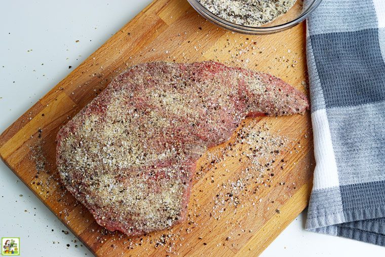 Uncooked tri-tip beef roast with rub spices on a wooden cutting board with black and white tea towel.