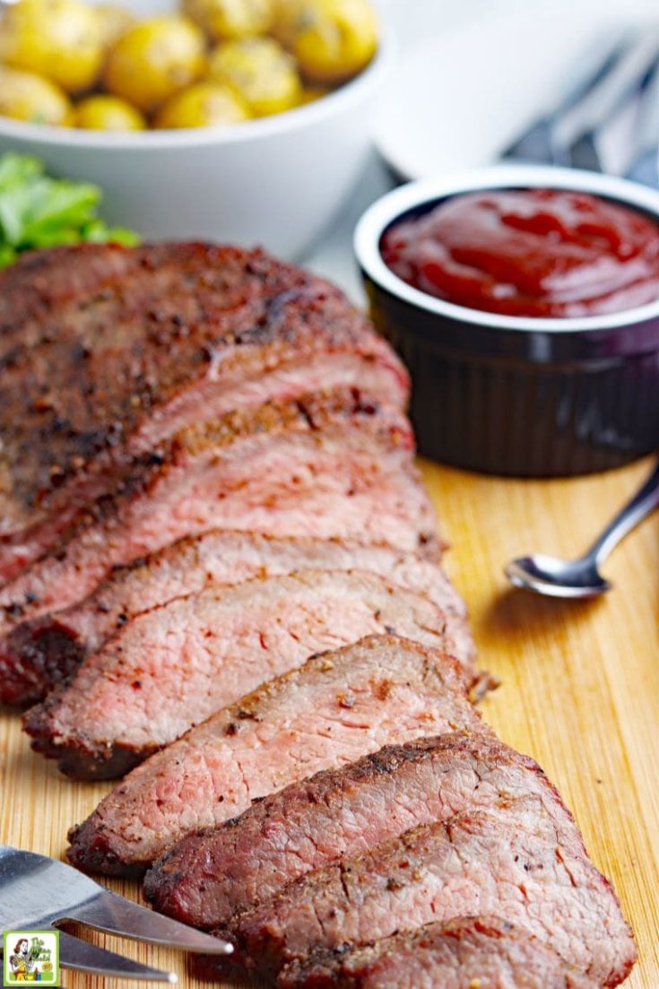 Close up of sliced smoke tri-tip beef roast with bowls of BBQ sauce and roasted potatoes.