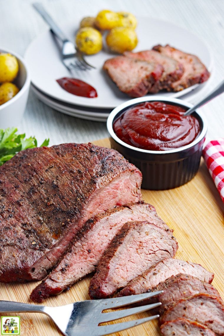 A tabletop with a plate of sliced smoked tri-tip roast, sliced roast on a cutting board, and a bowl of BBQ sauce.