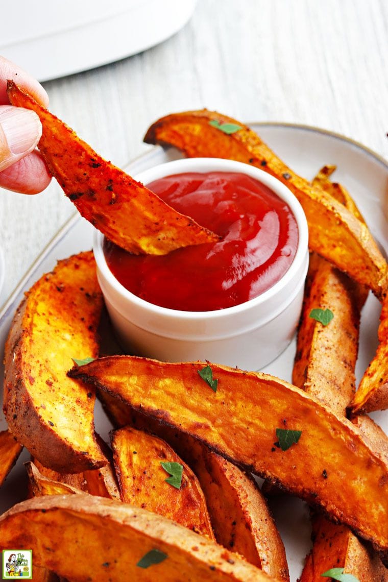Hand dipping a fried sweet potato wedge in a small pot of ketchup.