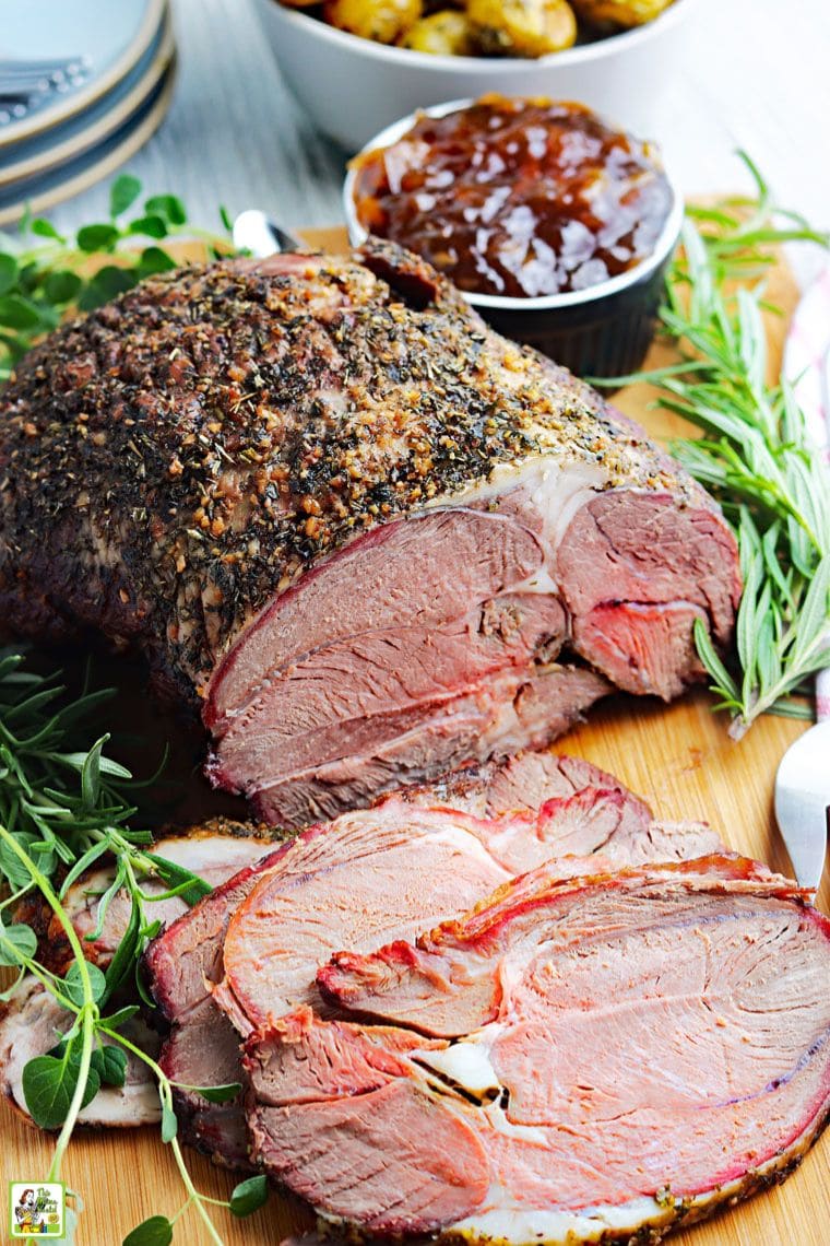 A sliced smoked leg of lamb roast on a cutting board with herbs, a pot of chutney sauce, and a bowl of potatoes.