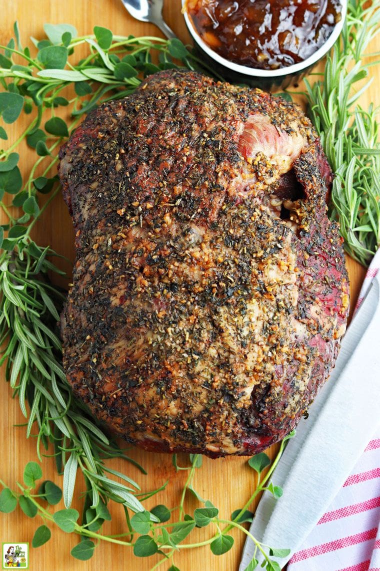 A cooked roasted leg of lamb before slicing surrounded by fresh herbs and cranberry sauce.